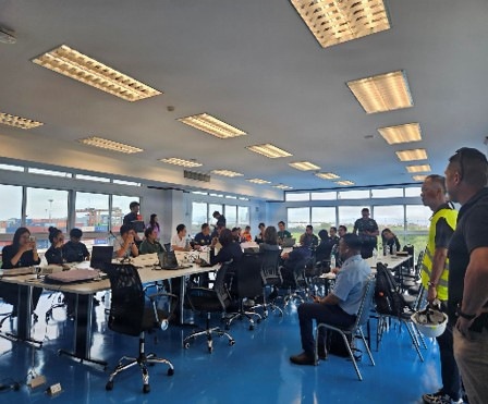 A large group of approximately 25-35 people are gathered in a modern conference or operations room with floor-to-ceiling windows overlooking what appears to be a port or industrial facility. Participants are seated at long tables with laptops and documents, suggesting a formal meeting, briefing, or training session. Some individuals are wearing high-visibility vests and uniforms, indicating a multi-agency or operational gathering.