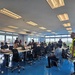 A large group of approximately 25-35 people are gathered in a modern conference or operations room with floor-to-ceiling windows overlooking what appears to be a port or industrial facility. Participants are seated at long tables with laptops and documents, suggesting a formal meeting, briefing, or training session. Some individuals are wearing high-visibility vests and uniforms, indicating a multi-agency or operational gathering.