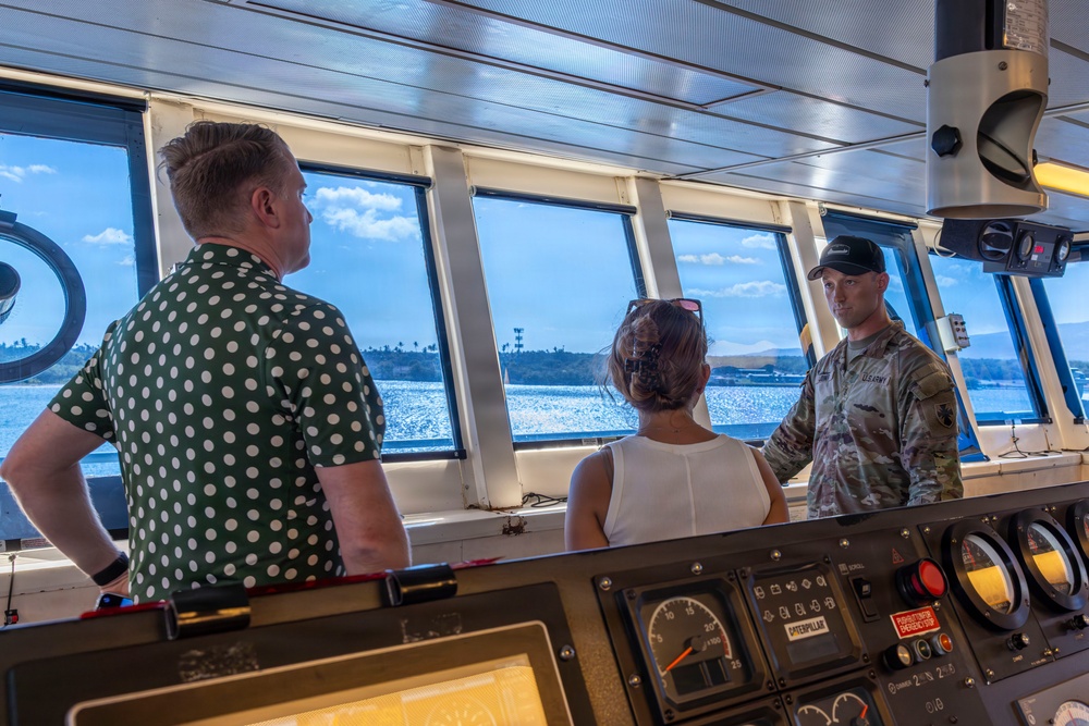 Three individuals are gathered on the bridge/wheelhouse of a vessel, with a U.S. Army soldier in multicam uniform appearing to brief or explain something to two civilians. The setting is a ship's command bridge featuring navigation and engine control instruments, with a waterfront and distant shoreline visible through the windows. The interaction suggests an official tour or briefing aboard a military or government vessel.