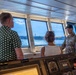 Three individuals are gathered on the bridge/wheelhouse of a vessel, with a U.S. Army soldier in multicam uniform appearing to brief or explain something to two civilians. The setting is a ship's command bridge featuring navigation and engine control instruments, with a waterfront and distant shoreline visible through the windows. The interaction suggests an official tour or briefing aboard a military or government vessel.