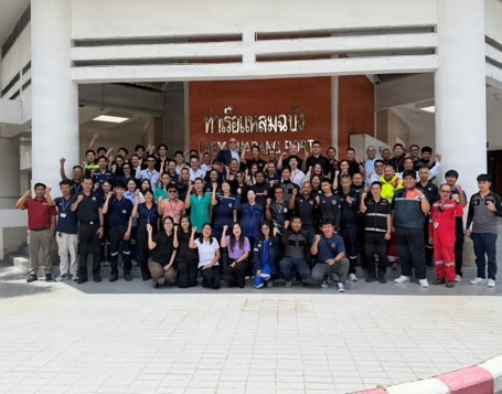 A large group of approximately 60-80 people are posing for a group photograph in front of the Laem Chabang Port building in Thailand. The participants are wearing various uniforms including dark workwear, high-visibility vests, and casual office attire, suggesting a multi-department or multi-organization gathering. Many individuals are raising their fists or giving thumbs-up gestures in a celebratory or solidarity pose.