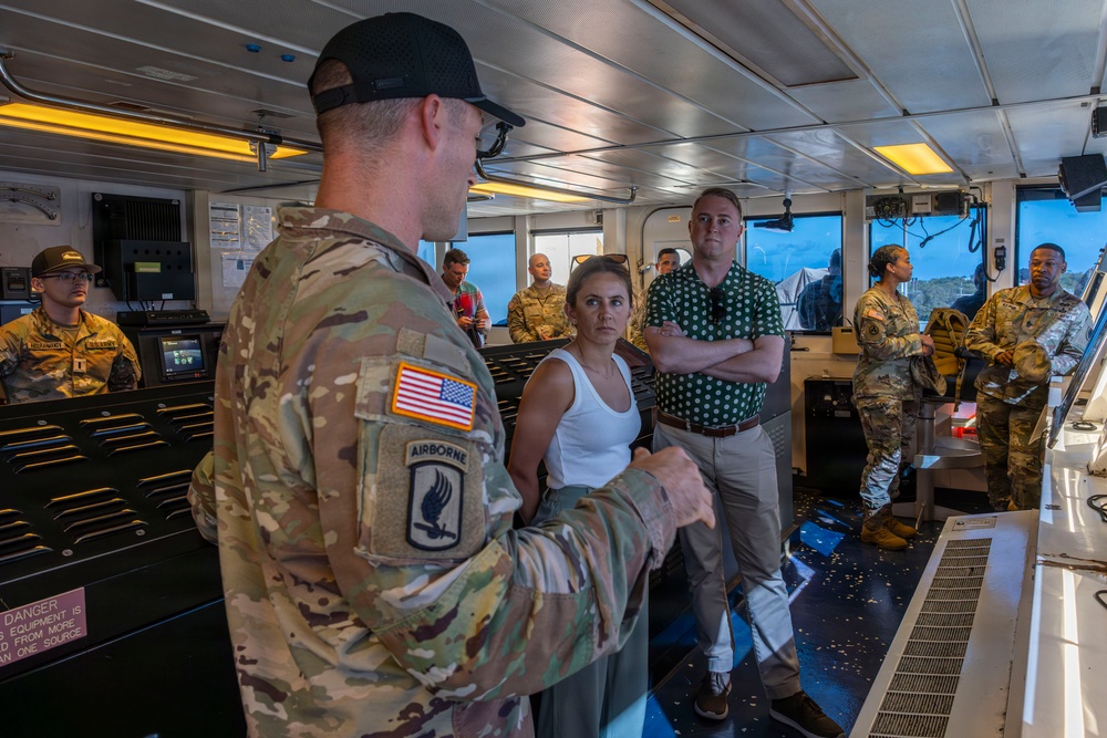 A U.S. Army soldier wearing 173rd Airborne Brigade insignia is briefing civilians and other military personnel aboard what appears to be a military vessel's bridge or command deck. The group includes both uniformed soldiers in OCP camouflage and civilians in casual attire, suggesting a tour or official visit. The setting appears to be an operational naval or watercraft environment with visible control consoles and equipment.