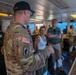 A U.S. Army soldier wearing 173rd Airborne Brigade insignia is briefing civilians and other military personnel aboard what appears to be a military vessel's bridge or command deck. The group includes both uniformed soldiers in OCP camouflage and civilians in casual attire, suggesting a tour or official visit. The setting appears to be an operational naval or watercraft environment with visible control consoles and equipment.