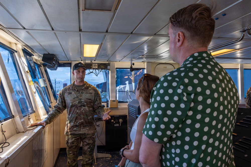 A U.S. Army soldier in camouflage uniform is briefing or giving a tour to two civilians inside what appears to be the bridge or pilothouse of a military or government vessel. The soldier is gesturing with his hand while speaking to a man in a green polka-dot shirt and a woman in a white top. The setting suggests an official tour or media engagement aboard a watercraft.