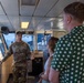 A U.S. Army soldier in camouflage uniform is briefing or giving a tour to two civilians inside what appears to be the bridge or pilothouse of a military or government vessel. The soldier is gesturing with his hand while speaking to a man in a green polka-dot shirt and a woman in a white top. The setting suggests an official tour or media engagement aboard a watercraft.
