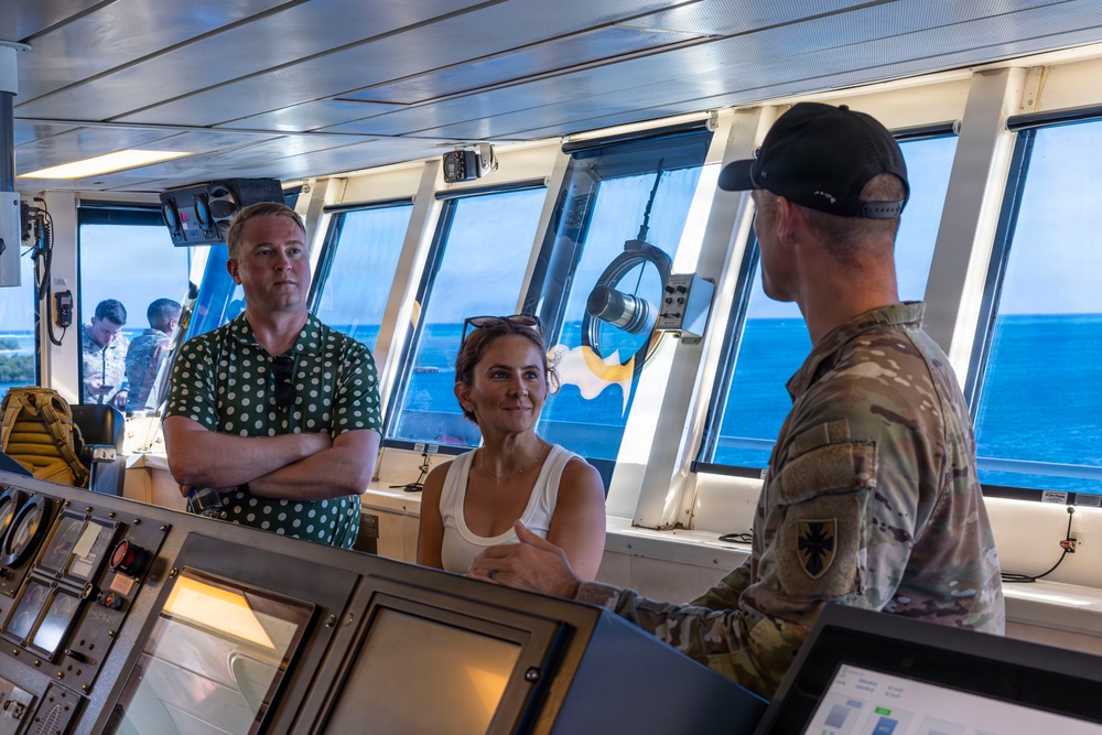 Three individuals are gathered on the bridge of a vessel, with a military personnel in camouflage uniform appearing to brief or explain something to two civilians. The setting is the ship's navigation bridge, identifiable by the control panels, navigation equipment, and large forward-facing windows overlooking open blue water. The interaction appears to be an official tour or briefing.