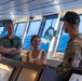 Three individuals are gathered on the bridge of a vessel, with a military personnel in camouflage uniform appearing to brief or explain something to two civilians. The setting is the ship's navigation bridge, identifiable by the control panels, navigation equipment, and large forward-facing windows overlooking open blue water. The interaction appears to be an official tour or briefing.