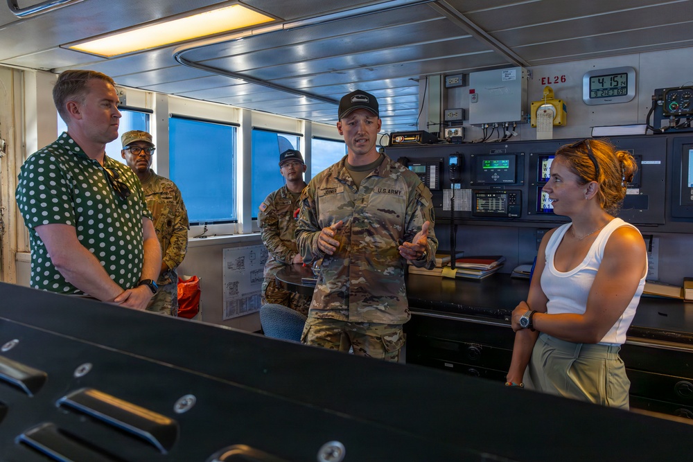 A U.S. Army soldier, identified by visible 'U.S. ARMY' and 'JONES' name tape on his uniform, appears to be briefing or explaining something to a small group aboard what appears to be a ship's bridge or control room. The group includes two civilians (one male in a green polka-dot shirt, one female in a white tank top) and at least two other military personnel in camouflage uniforms. The setting suggests an official tour or operational briefing aboard a military or government vessel.