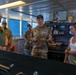 A U.S. Army soldier, identified by visible 'U.S. ARMY' and 'JONES' name tape on his uniform, appears to be briefing or explaining something to a small group aboard what appears to be a ship's bridge or control room. The group includes two civilians (one male in a green polka-dot shirt, one female in a white tank top) and at least two other military personnel in camouflage uniforms. The setting suggests an official tour or operational briefing aboard a military or government vessel.