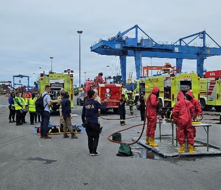 The image depicts an emergency response drill or incident at a commercial port facility, featuring multiple response teams including hazmat personnel in red protective suits, firefighters, and observers in high-visibility vests. Several yellow emergency response vehicles and a red fire truck are staged in the area, with large blue port cranes visible in the background. The scene suggests a coordinated multi-agency exercise involving hazardous materials response and decontamination procedures.