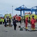 The image depicts an emergency response drill or incident at a commercial port facility, featuring multiple response teams including hazmat personnel in red protective suits, firefighters, and observers in high-visibility vests. Several yellow emergency response vehicles and a red fire truck are staged in the area, with large blue port cranes visible in the background. The scene suggests a coordinated multi-agency exercise involving hazardous materials response and decontamination procedures.