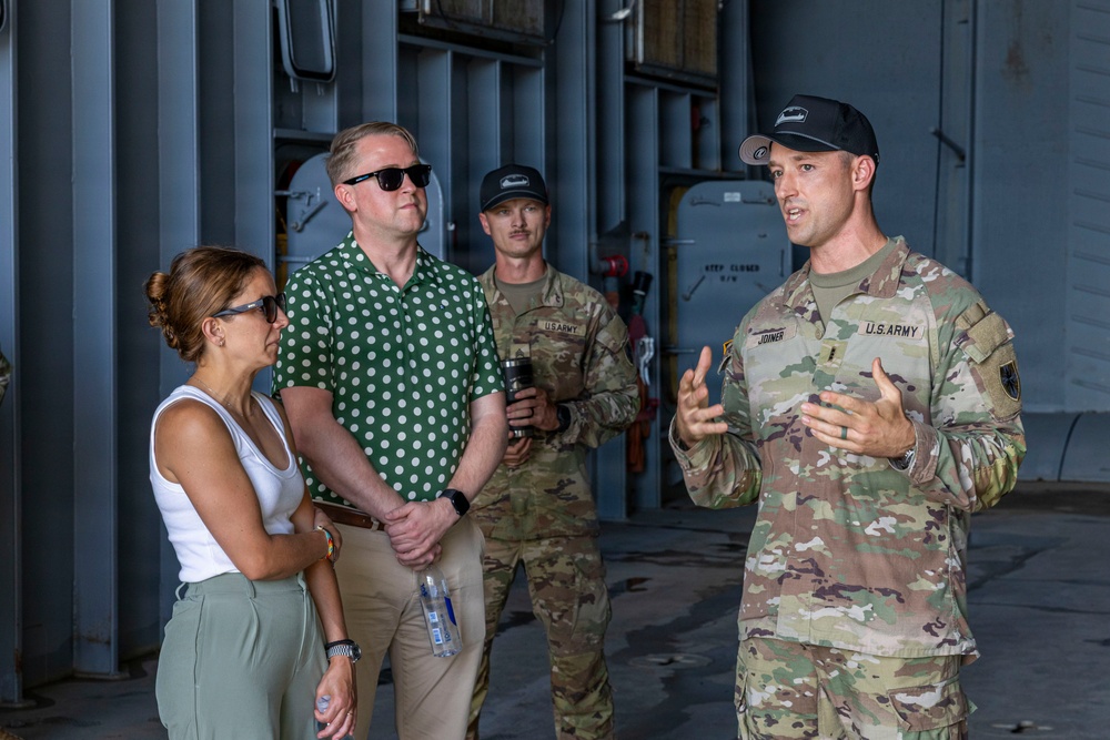 A U.S. Army soldier, identified by name tape as 'JOINER,' appears to be briefing or speaking to two civilians and another soldier in what looks like a military hangar or facility. The soldier in uniform is gesturing with his hands, suggesting an active explanation or presentation. Two civilians, a woman in a white top and a man in a green polka-dot shirt, are listening attentively.