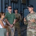 A U.S. Army soldier, identified by name tape as 'JOINER,' appears to be briefing or speaking to two civilians and another soldier in what looks like a military hangar or facility. The soldier in uniform is gesturing with his hands, suggesting an active explanation or presentation. Two civilians, a woman in a white top and a man in a green polka-dot shirt, are listening attentively.