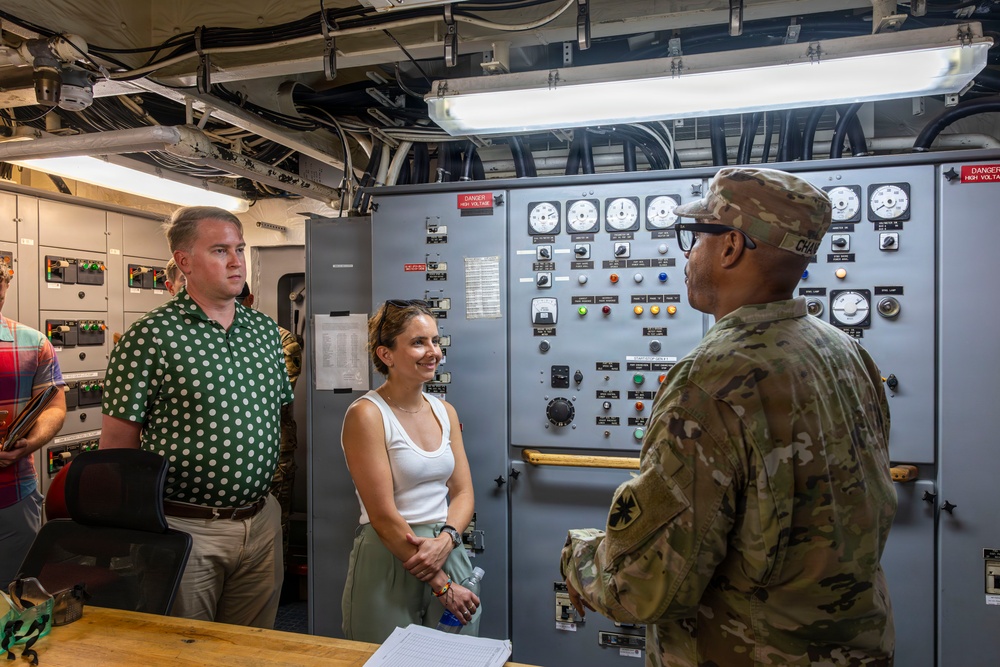 A uniformed military personnel member appears to be briefing or giving a tour to two civilians inside what appears to be a military vessel or facility control room. The setting features an industrial control panel with numerous gauges, switches, and indicator lights. The interaction appears formal yet relaxed, with the civilians attentively listening.