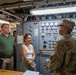 A uniformed military personnel member appears to be briefing or giving a tour to two civilians inside what appears to be a military vessel or facility control room. The setting features an industrial control panel with numerous gauges, switches, and indicator lights. The interaction appears formal yet relaxed, with the civilians attentively listening.