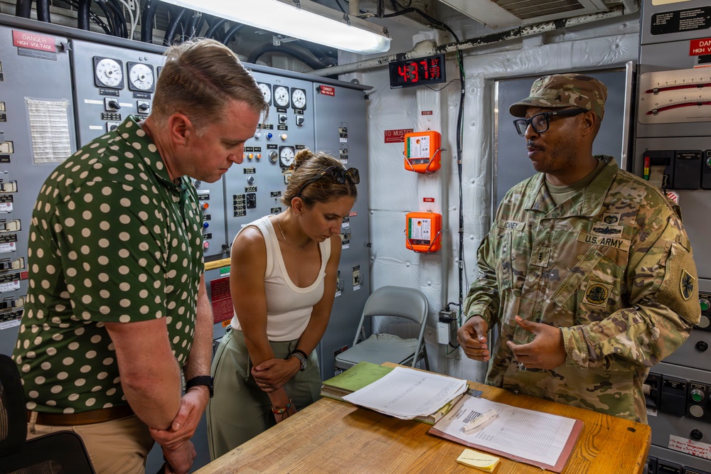 A U.S. Army soldier in multicam uniform is briefing or explaining something to two civilians inside what appears to be a military vessel or facility control room. The soldier, whose name tape reads 'MCONEY' and whose uniform bears 'U.S. ARMY' tape, is gesturing while speaking to a man in a green polka-dot shirt and a woman in a white top. Documents and papers are spread on a wooden table between them.