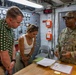 A U.S. Army soldier in multicam uniform is briefing or explaining something to two civilians inside what appears to be a military vessel or facility control room. The soldier, whose name tape reads 'MCONEY' and whose uniform bears 'U.S. ARMY' tape, is gesturing while speaking to a man in a green polka-dot shirt and a woman in a white top. Documents and papers are spread on a wooden table between them.