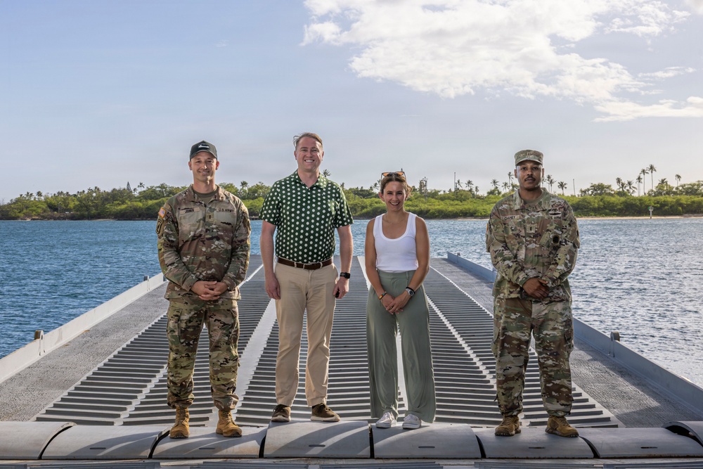 Four individuals are posing for a group photo on a floating dock or pier over open water, with a tropical island visible in the background. Two of the individuals are wearing U.S. Army camouflage uniforms, while the other two are in civilian attire. The setting appears to be a tropical coastal location, likely in the Pacific region.