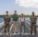 Four individuals are posing for a group photo on a floating dock or pier over open water, with a tropical island visible in the background. Two of the individuals are wearing U.S. Army camouflage uniforms, while the other two are in civilian attire. The setting appears to be a tropical coastal location, likely in the Pacific region.