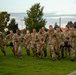 A large group of military personnel in OCP (Operational Camouflage Pattern) uniforms are running across a grass field, likely during a physical training exercise. Several soldiers in the front are carrying a litter (military stretcher) with a person on it, suggesting a casualty evacuation drill. The setting appears to be a military installation sports field during early morning or evening hours.