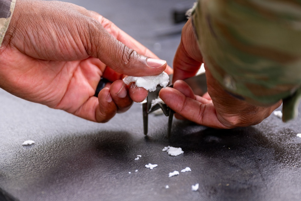 A person wearing military camouflage clothing is handling a white fibrous or powdery substance using a small tool, likely pliers or tweezers, over a dark surface. Small white particles are scattered on the dark surface below. The context suggests a maintenance, cleaning, or material-handling task.