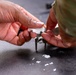 A person wearing military camouflage clothing is handling a white fibrous or powdery substance using a small tool, likely pliers or tweezers, over a dark surface. Small white particles are scattered on the dark surface below. The context suggests a maintenance, cleaning, or material-handling task.