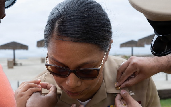 A military ceremony appears to be taking place outdoors near a beach area, where a person in a tan/khaki Navy uniform is having insignia or rank devices pinned onto their collar by two individuals. The central figure is looking down at their chest while two sets of hands work on attaching items to their uniform. This appears to be a promotion or pinning ceremony.