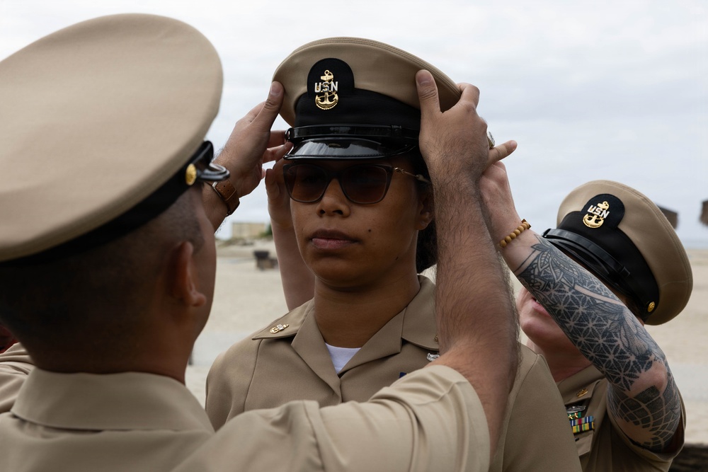 A U.S. Navy Chief Petty Officer pinning ceremony is taking place outdoors, where two individuals are placing a Chief Petty Officer combination cover (hat) onto a newly promoted sailor. The recipient is wearing tan Navy Service Uniform (NSU) and sunglasses, standing at attention while peers assist with the cover placement. This appears to be part of a Chief Petty Officer (CPO) initiation or frocking ceremony.