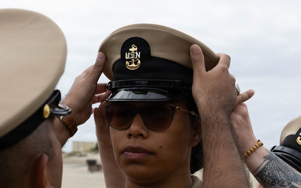 A U.S. Navy Chief Petty Officer pinning ceremony is taking place outdoors, where two individuals are placing a Chief Petty Officer combination cover (hat) onto a newly promoted sailor. The recipient is wearing tan Navy Service Uniform (NSU) and sunglasses, standing at attention while peers assist with the cover placement. This appears to be part of a Chief Petty Officer (CPO) initiation or frocking ceremony.