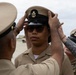 A U.S. Navy Chief Petty Officer pinning ceremony is taking place outdoors, where two individuals are placing a Chief Petty Officer combination cover (hat) onto a newly promoted sailor. The recipient is wearing tan Navy Service Uniform (NSU) and sunglasses, standing at attention while peers assist with the cover placement. This appears to be part of a Chief Petty Officer (CPO) initiation or frocking ceremony.