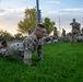 A group of U.S. Army soldiers in OCP (Operational Camouflage Pattern) uniforms are conducting physical training exercises on a grass field at dawn or dusk. The foreground features a soldier performing push-ups while wearing glasses, with numerous other soldiers doing the same exercise in the background. Several cadre or supervisory personnel are standing and observing the training session.