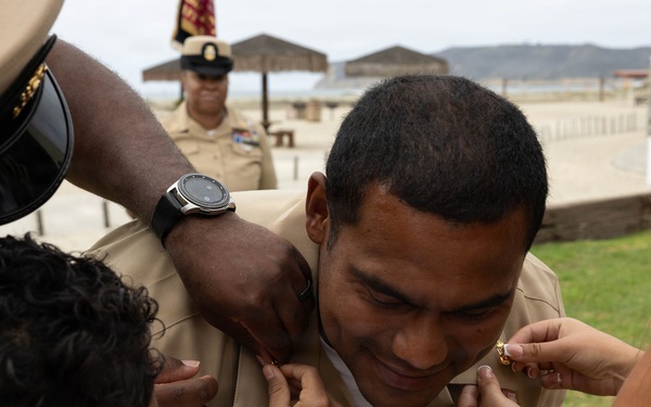 A military promotion ceremony is taking place outdoors near a beach or coastal area. Multiple individuals, including what appears to be family members and a senior officer, are pinning new rank insignia onto a smiling service member in a tan/khaki U.S. Navy or Marine Corps uniform. The subject appears pleased as several hands simultaneously attach pins to his collar.
