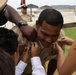 A military promotion ceremony is taking place outdoors near a beach or coastal area. Multiple individuals, including what appears to be family members and a senior officer, are pinning new rank insignia onto a smiling service member in a tan/khaki U.S. Navy or Marine Corps uniform. The subject appears pleased as several hands simultaneously attach pins to his collar.