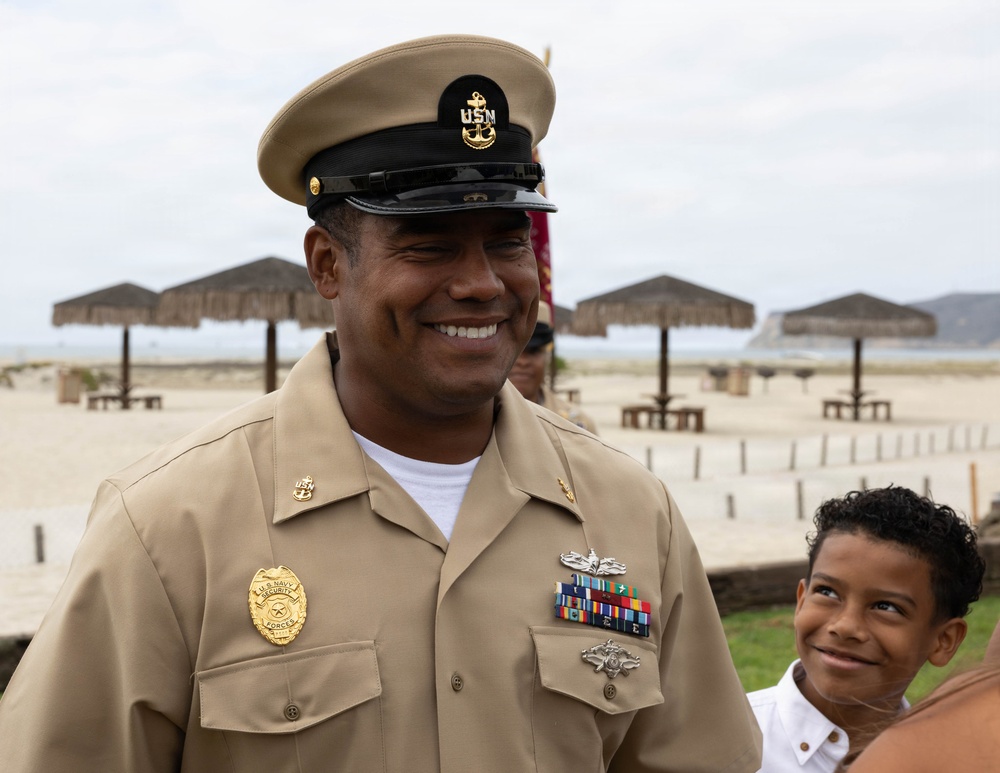 A U.S. Navy Chief Petty Officer in service khaki uniform is smiling at a beach location, likely during a ceremony or official event. He is wearing a combination cover with the USN anchor insignia, a Navy Security Forces badge, service ribbons, and qualification devices. A young boy in a white shirt is visible beside him, also smiling.