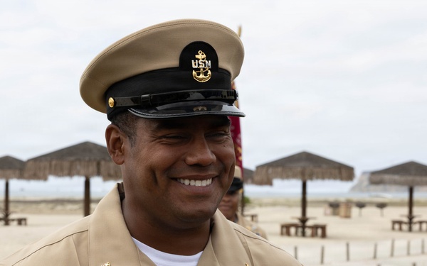 A U.S. Navy Chief Petty Officer in service khaki uniform is smiling at a beach location, likely during a ceremony or official event. He is wearing a combination cover with the USN anchor insignia, a Navy Security Forces badge, service ribbons, and qualification devices. A young boy in a white shirt is visible beside him, also smiling.