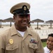 A U.S. Navy Chief Petty Officer in service khaki uniform is smiling at a beach location, likely during a ceremony or official event. He is wearing a combination cover with the USN anchor insignia, a Navy Security Forces badge, service ribbons, and qualification devices. A young boy in a white shirt is visible beside him, also smiling.