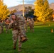 Military personnel in OCP (Operational Camouflage Pattern) uniforms are conducting a combat casualty carry drill on a grassy field, likely at a military installation. One soldier is performing a fireman's carry, transporting another soldier across the field toward an orange cone marker. The scene appears to take place during early morning or evening golden hour, with mountains and trees visible in the background.