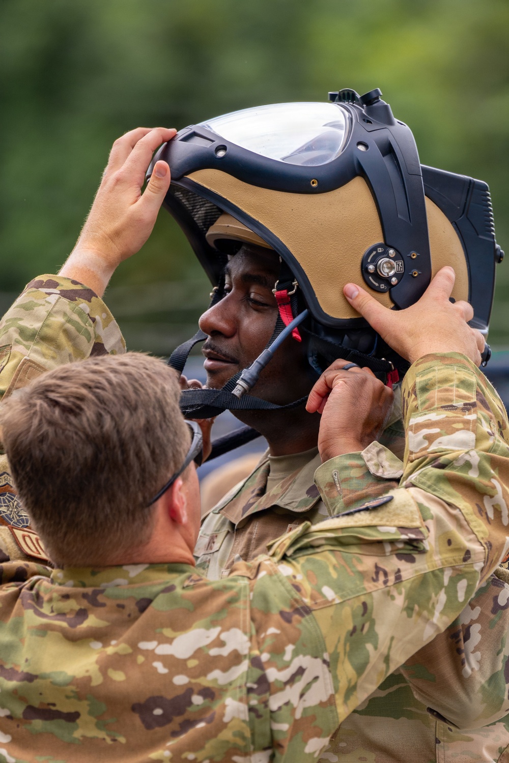 Two military personnel in MultiCam/OCP camouflage uniforms are shown, with one assisting the other in fitting a specialized helmet. The helmet appears to be a modern aircrew or EOD-style protective helmet with a large visor, tan/coyote brown shell, black frame, and integrated communication components. The scene appears to take place outdoors with green foliage visible in the background.