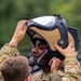 Two military personnel in MultiCam/OCP camouflage uniforms are shown, with one assisting the other in fitting a specialized helmet. The helmet appears to be a modern aircrew or EOD-style protective helmet with a large visor, tan/coyote brown shell, black frame, and integrated communication components. The scene appears to take place outdoors with green foliage visible in the background.