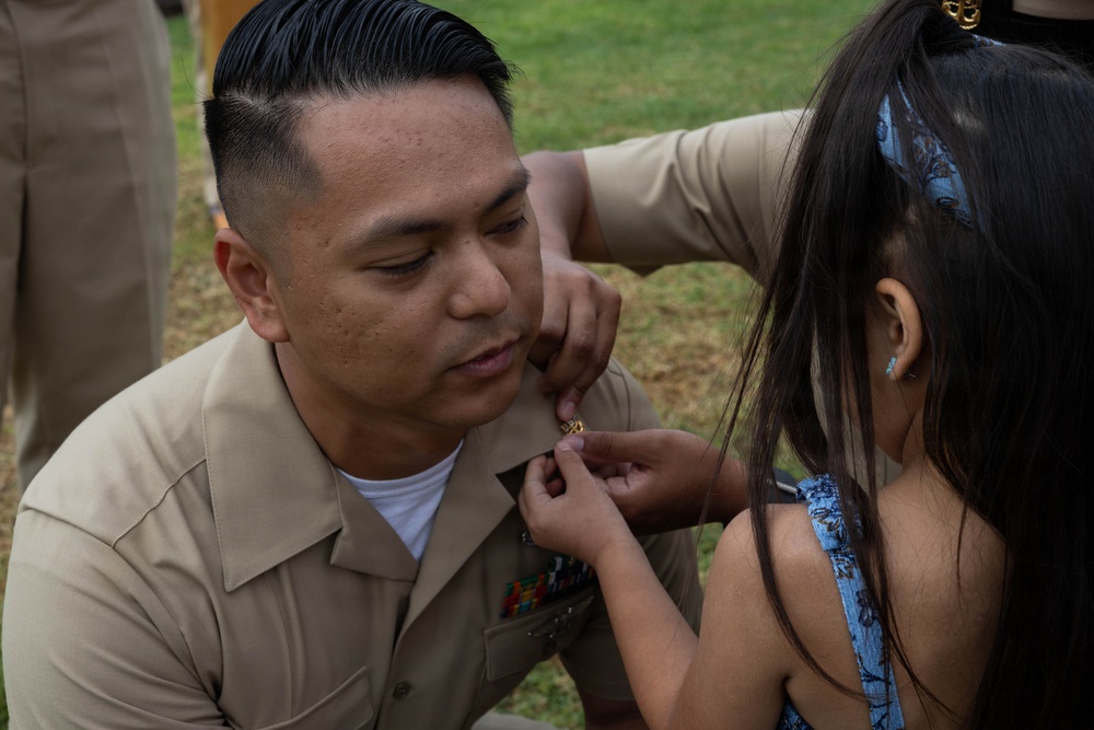 A U.S. Navy servicemember in tan/khaki uniform is being assisted by a young girl, likely a family member, in pinning an insignia or device onto his uniform collar. This appears to be a military promotion or advancement ceremony conducted outdoors on a grassy area. The moment captures an intimate ceremonial tradition where family members participate in the pinning.