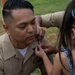 A U.S. Navy servicemember in tan/khaki uniform is being assisted by a young girl, likely a family member, in pinning an insignia or device onto his uniform collar. This appears to be a military promotion or advancement ceremony conducted outdoors on a grassy area. The moment captures an intimate ceremonial tradition where family members participate in the pinning.