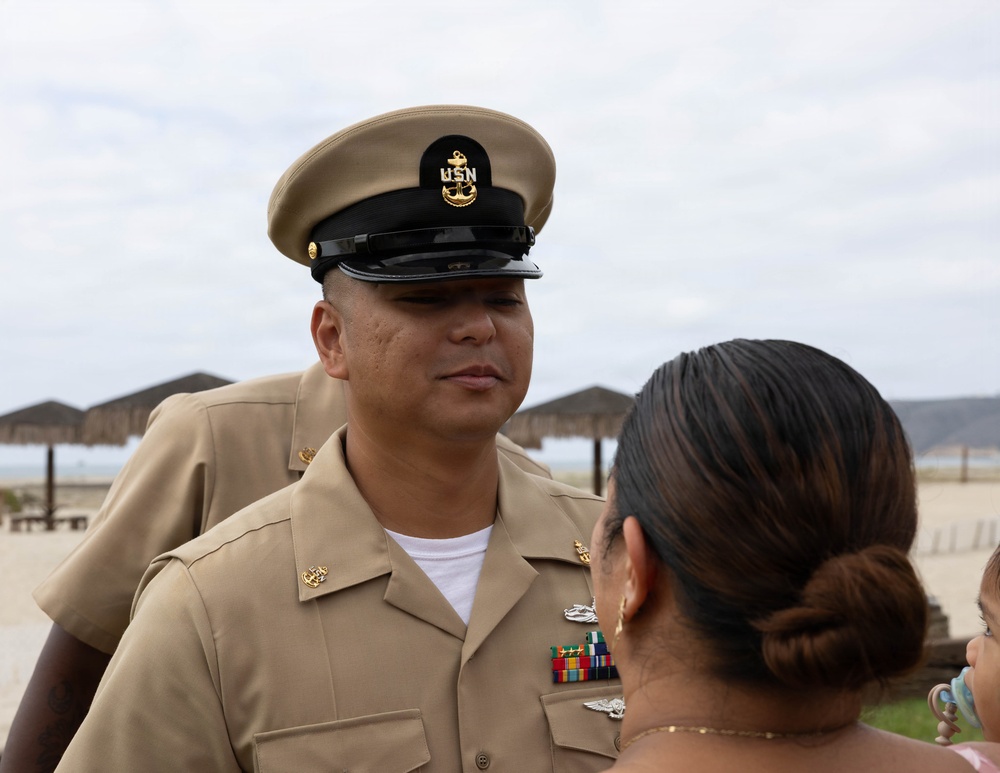 A United States Navy Chief Petty Officer in Service Khaki uniform is engaged in conversation with a civilian woman at what appears to be a beachside location. The sailor is wearing a combination cover with the USN anchor insignia and displays multiple service ribbons and warfare devices on his uniform. The setting appears to be an outdoor ceremony or informal gathering near a beach.