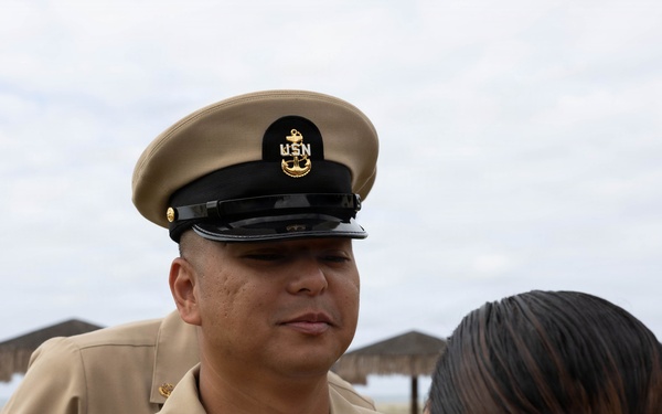 A United States Navy Chief Petty Officer in Service Khaki uniform is engaged in conversation with a civilian woman at what appears to be a beachside location. The sailor is wearing a combination cover with the USN anchor insignia and displays multiple service ribbons and warfare devices on his uniform. The setting appears to be an outdoor ceremony or informal gathering near a beach.
