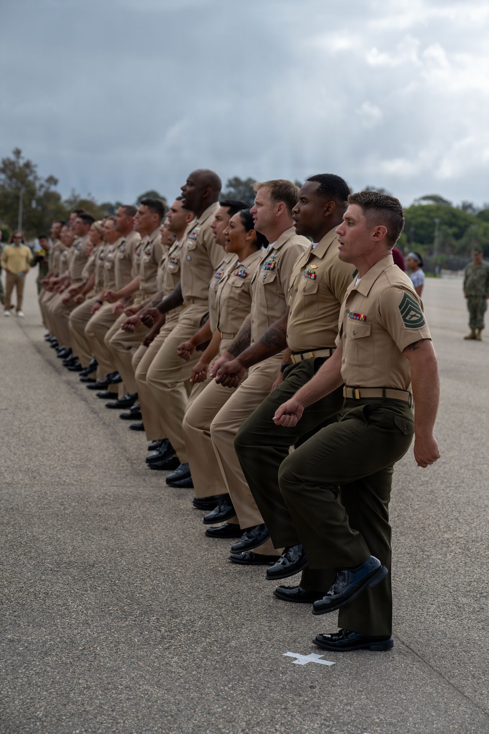 A formation of U.S. military personnel, primarily in tan/khaki service uniforms consistent with U.S. Marine Corps and Navy dress uniforms, are performing a synchronized movement during what appears to be a formal ceremony or parade. The group is standing in a diagonal line on a paved surface, with knees bent and arms positioned in a coordinated stance. The setting appears to be an outdoor military installation with overcast skies.