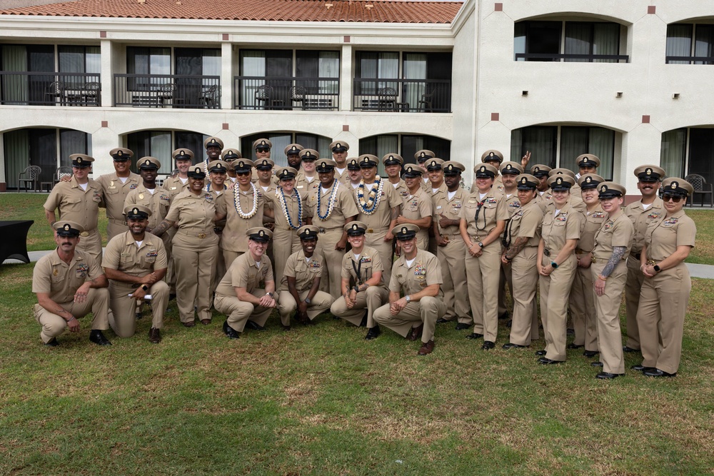 A large group of U.S. Navy personnel in tan/khaki service uniforms pose for a formal group photograph outdoors on a grass lawn. Several individuals in the center are wearing traditional Hawaiian lei garlands, suggesting a ceremony or celebration, likely at a naval installation in Hawaii. The group consists of both male and female sailors of various ranks, identifiable by their chief petty officer-style covers and insignia.