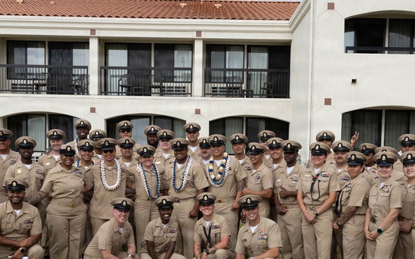 A large group of U.S. Navy personnel in tan/khaki service uniforms pose for a formal group photograph outdoors on a grass lawn. Several individuals in the center are wearing traditional Hawaiian lei garlands, suggesting a ceremony or celebration, likely at a naval installation in Hawaii. The group consists of both male and female sailors of various ranks, identifiable by their chief petty officer-style covers and insignia.