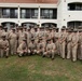 A large group of U.S. Navy personnel in tan/khaki service uniforms pose for a formal group photograph outdoors on a grass lawn. Several individuals in the center are wearing traditional Hawaiian lei garlands, suggesting a ceremony or celebration, likely at a naval installation in Hawaii. The group consists of both male and female sailors of various ranks, identifiable by their chief petty officer-style covers and insignia.