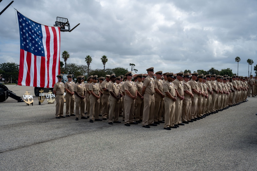 A large formation of U.S. Navy personnel in khaki service uniforms are standing at attention during a formal military ceremony. An American flag is prominently displayed on the left side, and USN anchor insignia decorations are visible at the base. The event appears to take place on a paved parade ground or tarmac, likely at a naval installation with palm trees visible in the background.