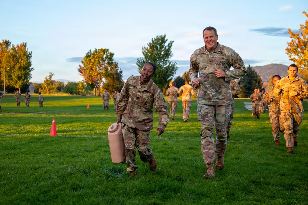A group of U.S. Army soldiers in OCP (Operational Camouflage Pattern) uniforms are participating in a physical training exercise on a grassy field during early morning or evening golden hour lighting. Two soldiers in the foreground — one carrying a large tan container — appear to be completing a fitness drill, with a senior officer running alongside them. Multiple other soldiers are visible in the background performing similar activities.