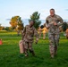 A group of U.S. Army soldiers in OCP (Operational Camouflage Pattern) uniforms are participating in a physical training exercise on a grassy field during early morning or evening golden hour lighting. Two soldiers in the foreground — one carrying a large tan container — appear to be completing a fitness drill, with a senior officer running alongside them. Multiple other soldiers are visible in the background performing similar activities.
